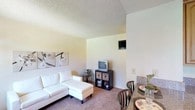 This single-floor unit offers a practical and comfortable layout across several interconnected spaces. The living and dining area is finished with wall-to-wall carpet and neutral-toned walls, creating a warm, understated atmosphere. A textured ceiling runs throughout the unit, and wall-mounted speakers are present in both the living room and bedroom. The dining area accommodates a table that flows naturally from the living space, with the two areas sharing an open connection.

The kitchen is well-equipped, featuring warm-toned oak cabinetry that extends to the upper walls, providing generous storage. Granite-style countertops offer ample workspace across an L-shaped layout, complete with a double-basin stainless steel sink, a coil-burner electric range, and space for small appliances. Light tile flooring grounds the kitchen area.

The bedroom is spacious with soft carpet underfoot and neutral walls. A large window allows natural light to fill the room, contributing to an airy feel. The textured ceiling continues here, maintaining consistency throughout.

The bathroom features tile flooring, a single-sink vanity with a large mirror framed by overhead lighting, and a combined tub and shower enclosure. The clean, light color palette keeps the space feeling bright and functional.