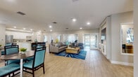 Located in Boynton Beach, Florida, this single-story community clubhouse at Boynton Place Circle offers a well-appointed gathering space designed to serve residents across multiple functional areas. The open-concept main lounge is expansive and bright, featuring light wood-look tile flooring, recessed lighting, and a coastal-inspired color palette of neutral grays and teal accents. A large sectional sofa, coffee table, and bold blue area rug anchor the seating area, while a wall-mounted flat-screen TV and built-in display shelving add a polished finishing touch. French doors at the far end of the lounge open to views of the outdoors, flooding the space with natural light. Adjacent to the lounge is a dining area with large windows dressed in decorative curtains, offering a bright and welcoming setting. The kitchen is sleek and modern, showcasing white cabinetry, granite countertops, a stainless steel refrigerator, and a central island with bar seating illuminated by pendant lighting. A dedicated fitness center provides a well-equipped workout space with strength-training machines, a weight bench, rubber flooring, and a wall-mounted TV, accented by bold geometric wall graphics. A compact business center nook with a built-in wood desk and computer workstation rounds out the amenities, making this clubhouse a comprehensive and versatile community hub.