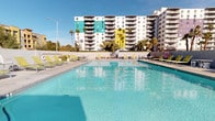 This outdoor amenity space centers around a generously sized swimming pool with crystal-clear turquoise water, framed by a wide concrete sun deck lined with bright yellow-green chaise lounges. The pool area offers ample room for relaxation and recreation, with easy entry access points and open sightlines to the surrounding urban skyline and lush palm trees. A covered patio lounge area adjacent to the pool provides a shaded retreat, featuring contemporary furnishings including a large curved sectional sofa with vibrant green cushions, a sleek coffee table atop a bold geometric area rug, and additional seating for dining or socializing. Floor-to-ceiling glass sliding doors connect the covered lounge to the pool deck, creating a seamless indoor-outdoor flow while flooding the space with natural light. The architectural design blends clean, modern lines with a resort-inspired aesthetic, complemented by a colorful multi-story building backdrop in teal and yellow accents. Together, these elements create a lively, well-appointed outdoor environment ideal for both casual gatherings and leisurely relaxation in a sunny, open-air setting.