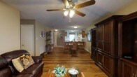 This single-story home showcases an open-concept floor plan that connects the living, dining, and kitchen areas in one continuous, welcoming space. Rich, dark hardwood flooring runs throughout, providing a warm and cohesive foundation across the main living areas.

The living room sits at the forefront of the space, furnished with a leather sofa and a wooden coffee table accented with decorative centerpieces. A ceiling fan with a built-in light fixture is centrally positioned overhead, complementing the textured ceiling. A large, dark wood entertainment armoire anchors one side of the room, offering ample storage and display space.

Flowing naturally into the dining area, a full-sized dining table with cushioned chairs provides a comfortable setting for meals. Decorative shelving and wall art add character along the neutral-toned walls.

The kitchen is visible at the far end of the space, featuring warm cabinetry, a window with valance treatments that invites natural light, and a counter with bar seating. The open sightlines throughout the layout make the home feel spacious and connected, ideal for both everyday living and entertaining.