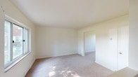 This two-floor residential space is designed with simplicity and functionality in mind, featuring a consistent neutral color palette of soft whites and warm beiges throughout. The flooring across both levels is wall-to-wall carpet in a light tan tone, providing a comfortable and cohesive feel from room to room.

The main level features a bright living area with a large picture window that frames views of a green, tree-lined exterior, allowing generous natural light to fill the space. A front entry door with a glazed panel provides additional light at the entry point. Floor-mounted HVAC vents are integrated throughout, ensuring climate comfort in each room.

The upper level is accessible via a staircase with a simple white handrail. The rooms on this floor are well-proportioned, with each featuring carpeted floors and crisp white walls. One room benefits from dual windows overlooking mature trees, while another is equipped with a ceiling fan and light fixture, complemented by two windows offering outdoor views. Additional rooms on this level are bright and open, with flush-mount ceiling fixtures providing overhead lighting.

Throughout the space, clean white baseboards frame each room, and the overall layout is open and practical. The neutral finishes and abundant natural light make this a versatile space well-suited for a variety of configurations and uses.
