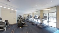 This contemporary fitness center offers a well-appointed workout environment designed to support a range of cardio and exercise routines. The space features a generous row of treadmills positioned along the window-facing wall, allowing users to exercise with natural light and outward views. Complementing the treadmills is a selection of elliptical cross-trainers and additional cardio machines, providing a comprehensive suite of equipment for all fitness levels. The interior is finished with a patterned, dark-toned carpet with accent detailing underfoot, adding both visual interest and durability suited for a high-traffic fitness environment. Overhead, sleek linear pendant lighting and recessed ceiling fixtures provide consistent, even illumination throughout the space. The walls are finished in a neutral warm tone, and a decorative stone accent wall serves as a design focal point, enhancing the overall aesthetic. Large windows fitted with roller shades allow for adjustable natural light, creating a bright and energizing atmosphere. Mirrored panels on one wall expand the sense of space visually while serving a functional purpose for form-checking during workouts. The layout is open and thoughtfully organized, ensuring ease of movement between equipment stations.