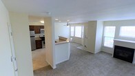 This two-floor space features a thoughtfully designed main level that integrates the kitchen, dining, and living areas into a cohesive open-concept layout. The kitchen is equipped with stainless steel appliances, including a refrigerator, stove, and microwave, complemented by rich dark wood cabinetry and granite-style countertops. A peninsula breakfast bar separates the kitchen from the living area, providing a functional transition between the two spaces. The living area is finished with plush neutral carpeting and benefits from abundant natural light streaming through multiple windows. A ceiling fan is centrally positioned overhead, enhancing airflow throughout the main level. The entry door is situated within a gently curved wall, adding a subtle architectural detail to the otherwise clean, neutral interior. Recessed lighting and flush-mount fixtures provide consistent illumination throughout. The overall color palette of soft whites and warm beiges creates a bright, welcoming ambiance. With two floors, the space offers additional room and flexibility beyond the main level.
