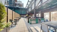 Located atop 95 Wall Street in the heart of New York City's Financial District, Roof Deck 2 is a thoughtfully designed outdoor amenity space that seamlessly blends leisure and fitness within an industrial-chic architectural framework. The deck is framed by dramatic black steel structural elements, floor-to-ceiling glass facades, and exposed brick walls, creating a bold urban aesthetic throughout.

The space is divided into distinct functional zones. A spacious paved terrace hosts multiple seating and dining areas furnished with modern grey tables and chairs, partially shaded by a retractable fabric pergola that provides comfort in varying weather conditions. Lush potted greenery adds a refreshing natural contrast to the hard surfaces. An outdoor kitchen station, accented with vibrant teal tile cladding and dark granite countertops, provides an ideal setup for entertaining and outdoor cooking.

Adjacent to the lounge area, a large artificial turf fitness lawn serves as an open-air workout zone, equipped with a power rack, punching bag, battle ropes, TRX suspension trainers, and cornhole boards for recreational use. Wooden privacy partitions define the perimeter while maintaining an open, airy feel. The surrounding NYC skyline provides a striking urban backdrop, making this roof deck a versatile and well-appointed amenity for residents.