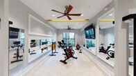 Located at 3938 Independence Blvd in Wilmington, NC, this single-floor fitness center offers a comprehensive workout facility with a thoughtfully designed layout divided into distinct functional zones. The space is finished with neutral gray walls, light-toned carpet with subtle yellow accents, and recessed lighting complemented by bold yellow-accented tray ceilings and coffered details that add a striking visual element overhead. Multiple ceiling fans are distributed throughout, ensuring comfort during workouts.

The main strength and cardio area features an array of Life Fitness and Freemotion equipment, including treadmills, cable machines, functional trainers, a punching bag, weight benches, and a full dumbbell rack. Large framed mirrors line the walls, enhancing the sense of space and allowing users to monitor their form. Flat-screen TVs are mounted throughout for entertainment.

A dedicated studio area accommodates cycling bikes, rowing machines, and a ballet barre, with its own wall-length mirror and mounted display screen. Colorful hand weights are neatly organized on a rack nearby. Sliding barn-style glass doors connect the studio to the main floor, maintaining an open, airy feel while providing separation between zones. Natural light filters in through multiple windows, adding to the clean, energizing atmosphere.