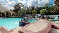 Located at 2220 Gloria Cir in Pensacola, Florida, this single-level outdoor amenity area offers a welcoming and well-appointed recreational space designed for relaxation and leisure. The centerpiece is a generously sized swimming pool with clear, blue water, complemented by an adjacent spa that provides a dedicated area for rest and hydrotherapy. The pool deck is finished with a decorative stamped concrete surface, providing both visual appeal and durability underfoot. Blue tile accents line the pool's edges, adding a polished aesthetic detail. A sturdy metal entry railing offers safe and convenient access into the water. Surrounding the pool, an expansive deck is furnished with multiple lounge chairs and seating areas, all arranged to take full advantage of the open sky and natural light. Mature palm trees and a backdrop of tall hardwoods frame the space, creating a lush, tropical atmosphere that is characteristic of the Florida Gulf Coast. A white privacy fence encloses the area, ensuring a sense of seclusion. Classic lamp posts add charm and provide lighting for extended use into the evening. The overall ambiance is bright, inviting, and resort-inspired, making this outdoor space a standout community amenity.