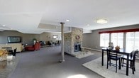 This single-floor space offers a generous, open layout designed for comfort and functionality. The room's most striking feature is a floor-to-ceiling natural stone fireplace positioned at the heart of the space, serving as a bold focal point that adds warmth and rustic character to the otherwise contemporary interior. A decorative column with a classic capital stands nearby, adding an architectural detail that complements the overall design. The flooring throughout is covered in neutral gray carpet, creating a cohesive and understated foundation for the space. The walls are finished in a warm taupe tone, contributing to an inviting and calm atmosphere. Recessed lighting and flush-mount ceiling fixtures provide even illumination throughout, while large windows along the perimeter allow generous natural light to fill the room. A seating area with a red accent chair and additional furnishings is visible toward one end, suggesting a lounge or gathering function. Subtle tray ceiling detailing adds dimension overhead. The overall ambiance is comfortable and welcoming, making it well-suited as a communal gathering or lounge space.