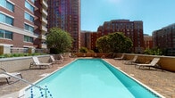 This single-floor residential unit is part of a prominent high-rise building complex distinguished by its classic brick exterior and urban skyline presence. The building's shared amenities are a standout feature, highlighted by a generously sized outdoor swimming pool surrounded by a paved brick terrace. The pool deck is lined with lounge chairs, offering residents a relaxing retreat beneath open skies, with mature trees and neighboring high-rise towers providing a dynamic urban backdrop.

Inside, the unit includes a bathroom finished in warm, neutral tones. The space features a wood-cabinet vanity with a white countertop basin, chrome fixtures, and a framed rectangular mirror accented by a lighted vanity bar overhead. A brushed gold electrical outlet and towel ring add refined, classic hardware details throughout. The toilet area is neatly tucked into its own alcove, complemented by a small decorative wall shelf. The overall palette is soft and cohesive, with cream-toned walls creating a calm, understated ambiance.

Together, the private interior spaces and resort-style outdoor amenities present a comfortable and functional living environment well-suited to urban residential life.