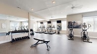Located at Carlton Manor in Sunbury, Ohio, this single-floor fitness center offers a generously sized, open workout environment designed to accommodate a full range of exercise needs. The space is finished with durable speckled rubber flooring throughout, providing both comfort and practicality for high-traffic use.

The main workout area is lined with an array of cardio equipment, including multiple treadmills and elliptical machines, all positioned to take advantage of the abundant natural light streaming through numerous windows fitted with blinds. Large wall-mounted mirrors amplify the sense of space and allow exercisers to monitor their form. Multiple flat-screen TVs are mounted on the walls, keeping members entertained during workouts, while ceiling fans ensure steady airflow throughout the room.

A dedicated strength training section houses a cable machine, a weight bench, and a well-stocked dumbbell rack with a full range of free weights. An adjacent flex area features open floor space and additional accessories such as stability balls, ideal for stretching and functional fitness. Glass-panel doors and interior windows connect this area to a secondary room visible from the main floor. Recessed lighting complements the natural light, creating a bright and welcoming atmosphere. The overall design is clean, neutral, and functional, making this fitness center a practical and inviting amenity.