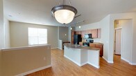 This single-floor space showcases an open-concept layout that seamlessly connects the kitchen and living areas. The kitchen is a standout feature, centered around a curved peninsula with granite countertops that provides both workspace and casual seating. Medium-toned wood cabinetry lines the kitchen walls, offering generous storage, while black appliances — including a refrigerator, electric range, and built-in microwave — lend a sleek, cohesive look. A track lighting system illuminates the kitchen area, complemented by a stylish pendant bowl light that hangs prominently over the peninsula, adding a decorative focal point. The flooring throughout is a warm hardwood-style material that extends continuously across the open space, contributing to a cohesive and inviting ambiance. A window with horizontal blinds allows natural light to filter into the living area, brightening the neutral gray-toned walls. White baseboards and trim provide clean contrast throughout. The open sightlines between the kitchen and adjacent living space make the layout feel spacious and functional, well-suited for both everyday living and entertaining.