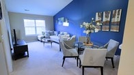 Located at 3998 Audrey Rae Ln in Howell, Michigan, this two-story home makes a striking first impression with its bold navy blue accent walls that anchor each room with a sense of depth and character. The main living area is generously proportioned, showcasing vaulted ceilings that enhance the sense of openness, while plush wall-to-wall carpeting adds warmth underfoot. A ceiling fan provides both comfort and style overhead. Natural light flows in through large windows, brightening the neutral tones that complement the vivid blue walls throughout. The living room flows seamlessly into the dining area, creating an open-concept layout ideal for everyday living and entertaining. The dining space features a glass-top round table nestled beside the bold accent wall, and connects directly to the kitchen via a convenient pass-through window — a practical architectural detail that keeps sightlines open while maintaining defined spaces. The kitchen is outfitted with white cabinetry and ample counter space. The bedroom continues the home's cohesive design aesthetic, featuring the signature navy accent wall paired with bright white walls and a window that offers outdoor views. The carpeted bedroom provides a calm, comfortable retreat. Throughout the home, the consistent use of color, texture, and thoughtful layout creates a cohesive and welcoming living environment.