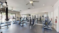 Located at 6640 East McDowell Road in Scottsdale, Arizona, this dedicated fitness center offers a comprehensive exercise environment within a compact, efficiently organized layout. The room features durable dark rubberized flooring throughout, providing a safe and functional surface suited for a wide range of workouts.

The cardio section is well-appointed with multiple treadmills, elliptical machines, and upright stationary bikes from brands including True Fitness, offering users a variety of aerobic training options. Strength training is equally well-represented, with a multi-station cable machine, a functional trainer with dual pulleys, selectorized weight machines, and a dedicated free weight rack stocked with a full set of dumbbells.

Full-length mirrors line one wall, visually expanding the space and allowing users to monitor their form during exercise. Large sliding glass windows along another wall flood the room with natural light and provide views of the surrounding outdoor landscape, contributing to an open and energizing atmosphere. Ceiling fans overhead ensure continuous air circulation for added comfort during workouts.

Recessed lighting and a clean, neutral color palette of white and light grey give the space a modern, professional aesthetic. The overall design prioritizes functionality and accessibility, making this fitness center a well-rounded amenity for those seeking a complete exercise facility.