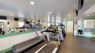 This fitness center is a comprehensively outfitted single-floor exercise facility designed to support a full range of workout routines. The space features a distinctive aesthetic defined by white beadboard ceilings with recessed lighting, and walls accented with a green horizontal stripe that adds a fresh, energetic character throughout.

The cardio area is generously stocked with multiple treadmills and elliptical trainers, each equipped with individual display screens. A wall of mirrors along one side visually expands the space and allows users to monitor their form during exercise. The rubber flooring throughout provides both durability and comfort underfoot.

A dedicated strength training section houses a multi-station cable machine along with benches and free weights, offering versatile resistance training options. Natural light filters in through several windows fitted with blinds, contributing to a well-lit and inviting atmosphere. Wall-mounted televisions are positioned throughout for entertainment during workouts.

A separate stretching and flexibility area includes yoga mats, a storage unit, and a wall-mounted TV, creating a quieter zone for cool-downs or low-impact exercise. A pedal exerciser and additional compact equipment round out this corner of the facility. The space is clean, organized, and well-maintained overall.