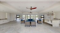 This expansive single-floor amenity space offers a seamless blend of indoor and outdoor living through its open, airy design. The layout is anchored by a generously sized granite-topped island at its center, complemented by sleek white cabinetry, a built-in stainless steel microwave, and an undermount sink — together forming a well-appointed kitchenette ideal for entertaining and gatherings.

The living area is furnished with comfortable wicker seating sets featuring deep blue cushions, arranged to encourage conversation and relaxation. Overhead, stylish ceiling fans with broad, sweeping blades ensure comfortable airflow throughout the space, while a decorative pendant light with an industrial-inspired cage design adds a refined focal point near the kitchen.

Polished concrete flooring runs throughout the entire space, providing a clean, modern aesthetic that is both durable and easy to maintain. Crisp white walls amplify the brightness, further enhanced by abundant natural light streaming in through wide, floor-to-ceiling black iron gated openings that connect the interior to the surrounding outdoor environment. These large gated walls offer sweeping views of the lush greenery and neighboring outdoor amenities, creating a naturally ventilated, resort-style atmosphere. The overall ambiance is modern, bright, and welcoming — well-suited for community events and everyday leisure.