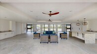 This expansive single-floor amenity space offers a seamless blend of indoor and outdoor living through its open, airy design. The layout is anchored by a generously sized granite-topped island at its center, complemented by sleek white cabinetry, a built-in stainless steel microwave, and an undermount sink — together forming a well-appointed kitchenette ideal for entertaining and gatherings.

The living area is furnished with comfortable wicker seating sets featuring deep blue cushions, arranged to encourage conversation and relaxation. Overhead, stylish ceiling fans with broad, sweeping blades ensure comfortable airflow throughout the space, while a decorative pendant light with an industrial-inspired cage design adds a refined focal point near the kitchen.

Polished concrete flooring runs throughout the entire space, providing a clean, modern aesthetic that is both durable and easy to maintain. Crisp white walls amplify the brightness, further enhanced by abundant natural light streaming in through wide, floor-to-ceiling black iron gated openings that connect the interior to the surrounding outdoor environment. These large gated walls offer sweeping views of the lush greenery and neighboring outdoor amenities, creating a naturally ventilated, resort-style atmosphere. The overall ambiance is modern, bright, and welcoming — well-suited for community events and everyday leisure.