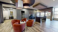 Located at 2525 W Tennessee St in Tallahassee, FL, this thoughtfully designed amenity center spans a single floor and comprises seven distinct functional areas. The welcoming main lounge immediately sets the tone with its striking sculptural ceiling detail featuring bold circular cutouts, oversized drum pendant lights, and plush terracotta swivel chairs arranged around a central coffee table. Wood-look tile flooring flows seamlessly throughout the space, lending a warm and cohesive aesthetic. A sleek bar-height counter with pendant lighting serves as a reception or social hub adjacent to the lounge. A dedicated business center offers multiple workstations equipped with desktop computers and privacy dividers, accented by contemporary artwork. Two study or small meeting rooms feature round tables, wall-mounted flat-screen TVs, large whiteboards, and ample natural light from oversized windows with outdoor views. A formal conference room provides a more polished setting, complete with a dark wood table, upholstered seating, elegant pendant fixtures, and curated wall art beneath a deep burgundy ceiling. A bright retail convenience area features floor-to-ceiling windows, ceiling fans, and well-stocked shelving. Throughout the space, colorful accent walls, modern lighting, and carefully chosen furnishings contribute to a vibrant yet functional atmosphere.