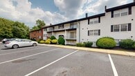 Located at 1803 Cobourg Ct in Parkville, Maryland, this parking lot serves a well-established residential community composed of multiple multi-story buildings. The complex features a mix of classic brick and white-painted exteriors, with black shutters and balconies adding architectural character to the facades. The parking area is expansive, offering numerous clearly marked, striped parking spaces laid out in an organized, accessible configuration. The lot is well-maintained with clean paving and defined curbing, providing straightforward ingress and egress throughout the property. Surrounding the buildings, neatly trimmed shrubbery, ornamental bushes, and mature trees contribute to a pleasant, green environment. The lush tree canopy visible along the perimeter of the community adds a sense of natural separation from the surrounding neighborhood. The community's layout, with parking centrally positioned between the residential buildings, ensures convenient access for residents. The grounds reflect a well-cared-for community setting, with landscaping maintained along building foundations and common walkways. Overall, this parking facility provides practical, ample vehicle accommodation within a charming, verdant residential community in Parkville, MD.