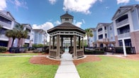 Located at 1001 Bear Island Rd in Summerville, South Carolina, this outdoor community space offers a welcoming and well-maintained amenity area designed for leisure and relaxation. At the heart of the space stands a classic octagonal gazebo with a distinctive multi-tiered roof topped by a decorative finial. Supported by sturdy columns with lattice detailing, the gazebo provides a shaded retreat surrounded by lush, manicured landscaping. A concrete walkway leads directly to the gazebo, flanked by neatly mulched garden beds and green lawns that enhance the inviting atmosphere. Beyond the gazebo, a wrought iron fence encloses a community pool area, visible in the background alongside a pergola structure. Mature palm trees and other established plantings add a touch of Southern charm and tropical warmth to the environment. The surrounding residential buildings, with their blue-grey siding and balconied facades, frame the space and provide a sense of community. Under the open South Carolina sky, this outdoor amenity area strikes a balance between functional design and aesthetic appeal, offering residents a pleasant gathering and recreational destination year-round.