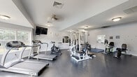 This fitness center at 454 Crestmont Ct in Copley, Ohio, offers a well-organized and fully equipped exercise facility spread across a single floor. The main workout area features durable dark rubber flooring throughout, providing a practical and resilient surface suited for a wide range of fitness activities. The space is bright and welcoming, benefiting from abundant natural light that streams in through a bank of large windows along one wall, offering pleasant outdoor views of surrounding greenery.

The cardio section is well-appointed with multiple treadmills, spinning bikes, an elliptical, and a recumbent bike, catering to varied cardiovascular training preferences. A wall-mounted flat-screen television accompanies the cardio area for entertainment during workouts. The strength training zone features a multi-station cable machine, a weight bench, stability balls, and a foldable exercise mat for floor work. Wall-mounted mirrors are positioned strategically to assist with form and to visually expand the space.

Ceiling fans and recessed lighting ensure comfort and adequate illumination throughout the room. The angled ceiling adds architectural character to the interior. A connecting hallway leads to clearly marked men's restroom facilities, adding convenience to the overall layout. The space presents a clean, neutral aesthetic with white walls and a functional, no-frills design focused on delivering a comprehensive workout experience.