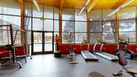 Located at 5400 N Nevada Ave in Colorado Springs, Colorado, this fitness center offers a thoughtfully designed workout environment that balances function with an inviting architectural character. The single-floor space is defined by soaring vaulted ceilings finished with warm wood planking and exposed structural beams, lending the room a distinctive and welcoming aesthetic. Floor-to-ceiling windows line the walls, flooding the interior with abundant natural light and providing open views of the surrounding outdoor area, including a pool and mature trees. Bold red accent walls create an energizing atmosphere complemented by durable rubber flooring throughout. The gym is comprehensively equipped to serve a wide range of fitness goals. Cardio equipment includes multiple treadmills, elliptical trainers, and stationary and spin bikes. Strength training is well represented with a Smith machine, weight bench, free weights, cable machines, and a functional training dummy. Additional benches and rowing-style equipment round out the offering. A glass-paned interior door connects to an adjacent lounge or common area, enhancing the overall accessibility of the facility. Double exterior doors open directly to the outdoor pool area, making this fitness center a seamlessly integrated part of a larger recreational complex.