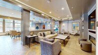 Located at 100 Sobel Dr E in Old Bridge, NJ, this thoughtfully designed amenity suite spans a generous single floor and offers residents a comprehensive selection of upscale shared spaces. The welcoming lounge area sets a refined tone with its tray ceilings, warm recessed and pendant lighting with gold accents, and a sophisticated mix of neutral-toned sofas and armchairs arranged around a central coffee table. A patterned geometric tile floor adds visual interest throughout the common areas. Adjacent to the lounge is a bright dining room bathed in natural light from large windows with garden views, furnished with multiple seating arrangements and complemented by a contemporary multi-arm chandelier. The fully equipped kitchen features crisp white cabinetry, under-cabinet LED lighting, white quartz countertops, gold hardware accents, and stainless steel appliances, ideal for resident gatherings and events. The expansive fitness center is outfitted with a full array of cardio machines, including treadmills and ellipticals, as well as comprehensive strength training equipment, all set on durable flooring beneath bright linear lighting and large windows. A dedicated studio space with floor-to-ceiling mirrors, motivational wall graphics, and light hardwood-style flooring provides an ideal setting for yoga, stretching, or group classes. Transitional corridor spaces with built-in shelving and accent seating complete this polished amenity floor.