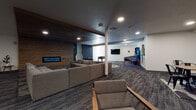 This contemporary community amenity space is thoughtfully designed to offer a variety of social and recreational experiences within a single, open floor plan. The space is anchored by a stylish lounge area featuring a large sectional sofa arranged around a wood-paneled accent wall with a sleek, wall-mounted electric fireplace displaying a vivid blue flame — creating a warm and inviting focal point. A bold geometric-patterned area rug adds visual interest underfoot, complementing the space's modern aesthetic.

Adjacent to the lounge, a dedicated game room area houses classic arcade cabinets, a pinball machine, and a foosball table, all set against clean white walls with recessed lighting above. A wall-mounted flat-screen television adds to the entertainment options in this zone.

A central gathering area with an industrial-style dining table and metal chairs provides a functional space for group activities or casual meetings. Additional seating is distributed throughout, including modern accent chairs with wooden armrests atop oval braided rugs, offering flexible and comfortable spots for relaxation.

The space features consistent dark-toned, multi-tonal carpeting throughout, recessed ceiling lighting, and a mix of dark wood and white ceiling finishes that define distinct zones. A color-blocked blue accent wall with a retro graphic mural adds a playful, energetic design element, reinforcing the space's vibrant and social character.