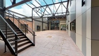 This striking covered courtyard showcases a compelling fusion of industrial heritage and contemporary design. At its most dramatic feature, an expansive steel truss framework supports a transparent skylight roof that floods the space with natural daylight, framing open views of the sky above. The generous courtyard floor is finished in stamped concrete with a warm, earthy tone, lending both durability and visual texture underfoot.

The surrounding walls present a layered mix of materials: original exposed brick on one side speaks to the building's historic character, while modern panel cladding in neutral tones defines the newer construction. Steel-framed glass doors and windows punctuate the perimeter, connecting the courtyard to interior corridors and office or workspace areas visible beyond.

A metal staircase with clean, modern railings rises along one side of the courtyard, providing multi-level access and contributing to the industrial aesthetic. A bank of mailboxes is mounted along one wall, indicating a multi-tenant use for the building. Seating benches and a teal wire chair are positioned throughout, creating informal gathering areas. A bicycle rack tucked near a painted brick wall offers practical amenities for occupants.

The overall ambiance is open, airy, and functional — a well-designed communal hub that bridges the building's historic roots with modern architectural sensibilities.