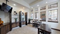This single-story space in Houston, TX (77070) is designed as a well-appointed communal or amenity area, offering a thoughtfully organized layout across several functional zones. The main lounge area features elevated tray ceilings with recessed lighting, light-toned carpet flooring, and an expansive bank of large, multi-pane windows that flood the interior with natural light and provide views of the surrounding outdoor areas, including what appears to be a pool. Dark espresso-finished open shelving units with lower cabinetry line one wall, serving as both decorative and functional storage, while a ceiling-mounted flat-screen television adds a media component to the space. Dark wood dining and high-top tables with matching stools provide multiple casual seating arrangements throughout. A separate kitchenette area is accessible via a tiled entryway framed by stone-clad columns. This room features L-shaped dark cabinetry with granite-style countertops, a sink, a coffee station, a microwave, a compact refrigerator, and a wall-mounted television. The warm ceramic tile flooring and a framed mirror complete the practical yet polished aesthetic of this service area, making the overall space well-suited for gathering, working, or relaxing.