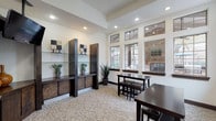 This single-story space in Houston, TX (77070) is designed as a well-appointed communal or amenity area, offering a thoughtfully organized layout across several functional zones. The main lounge area features elevated tray ceilings with recessed lighting, light-toned carpet flooring, and an expansive bank of large, multi-pane windows that flood the interior with natural light and provide views of the surrounding outdoor areas, including what appears to be a pool. Dark espresso-finished open shelving units with lower cabinetry line one wall, serving as both decorative and functional storage, while a ceiling-mounted flat-screen television adds a media component to the space. Dark wood dining and high-top tables with matching stools provide multiple casual seating arrangements throughout. A separate kitchenette area is accessible via a tiled entryway framed by stone-clad columns. This room features L-shaped dark cabinetry with granite-style countertops, a sink, a coffee station, a microwave, a compact refrigerator, and a wall-mounted television. The warm ceramic tile flooring and a framed mirror complete the practical yet polished aesthetic of this service area, making the overall space well-suited for gathering, working, or relaxing.