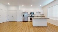 This single-floor unit is designed with a clean, modern aesthetic, showcasing white walls, recessed lighting, and light-toned hardwood flooring that flows through the main living areas. The open-concept layout connects the living space to a well-appointed kitchen, which features crisp white cabinetry with sleek bar-pull hardware, granite countertops, a central island, and a full suite of stainless steel appliances including a gas range, built-in microwave, dishwasher, and a side-by-side refrigerator with an in-door water and ice dispenser. Large windows allow natural light to fill the living area, enhancing the bright, airy atmosphere. The bedroom is carpeted for comfort and includes a tray ceiling detail, a second window, and sliding glass doors that open onto a private balcony with outdoor views. A dedicated closet area accommodates a stacked GE washer and dryer unit alongside wire shelving for additional storage. The bathroom continues the unit's clean design with a full soaking tub and shower combination, white surround, chrome fixtures, and granite-topped vanity. The overall ambiance is contemporary and move-in ready.