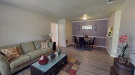 Located at 701 N Indiana Ave in Lubbock, Texas, this single-story home offers a welcoming and functional layout designed for comfortable everyday living. The open-concept living and dining area serves as the heart of the home, creating a seamless flow between spaces. The living area features hardwood-style flooring, a textured ceiling, and soft neutral wall tones that lend a warm and inviting atmosphere. A comfortable seating arrangement centers around a glass-top coffee table, making it an ideal space for relaxing or entertaining guests. The dining area is distinguished by a rich accent wall in a deep taupe-purple hue, complemented by a chair rail molding detail that adds a touch of architectural character. Wall sconces flank a small window, providing both ambient lighting and decorative interest. The dining space comfortably accommodates a full table and chair set, making it well-suited for family meals or gatherings. Hardwood-style flooring extends throughout, tying the living and dining spaces together cohesively. A hallway visible from the living area provides access to additional rooms, suggesting a thoughtful and practical floor plan. Overall, this home presents a clean, move-in-ready aesthetic with classic design details throughout.