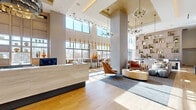 Located at 2501 S Ninth Road in Arlington, Virginia, this leasing office is a showcase of contemporary design and refined hospitality. The single-floor space makes a striking first impression with its soaring double-height ceilings and expansive floor-to-ceiling windows that flood the interior with natural light, offering views of the surrounding landscape. Warm wood-tone flooring flows throughout, anchoring the open, airy layout.

At the heart of the space stands an elegant reception desk finished in light wood and stone-toned materials, topped by a sculptural, layered pendant chandelier that serves as a focal point. Nearby, a dramatic large-scale abstract mural in soft blues, whites, and golds spans an entire wall, complemented by a built-in upholstered bench with decorative throw pillows — creating an inviting moment of pause.

The lounge area features a generous sectional sofa, accent chairs in warm cognac tones, and a floor-to-ceiling modular shelving wall displaying curated décor and greenery. A bar-height counter runs along the window wall, illuminated by a row of slender gold pendant lights, providing functional workspace with outdoor views. The overall ambiance is polished yet welcoming, balancing modern minimalism with warm, thoughtful details throughout.