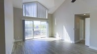 This single-floor space makes a striking first impression with its dramatic vaulted ceilings that soar to impressive heights, creating an open and airy atmosphere throughout. The main living area is anchored by wide-plank hardwood-style flooring in a cool gray tone, lending a modern and clean aesthetic to the space.

Large sliding glass doors span a generous portion of the exterior wall, inviting abundant natural light deep into the interior and offering views of the surrounding landscape, including lush greenery and neighboring rooftops. The doors also provide direct access to an outdoor area, seamlessly blending indoor and outdoor living.

High above the main living area, an elevated loft-style balcony railing is visible beneath the peak of the vaulted ceiling, adding architectural depth and visual interest to the space. Vertical blinds frame the upper clerestory windows, offering light control while maintaining the room's bright, open character.

The neutral, warm-gray wall tones complement the flooring and trim, creating a cohesive and versatile palette. A baseboard heater along the exterior wall ensures year-round comfort. The overall ambiance is one of spaciousness, light, and modern simplicity — a well-designed space that balances architectural character with functional living.