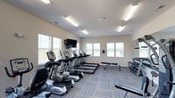 The fitness center at 1010 Fairway Village in Whitsett, North Carolina offers a well-equipped, single-floor exercise facility designed to support a variety of workout routines. The space is outfitted with a row of True-brand treadmills and elliptical machines, providing ample cardio options for residents. A dedicated free weight section along the far wall includes a multi-tiered dumbbell rack, while a weight bench situated centrally in the room accommodates strength training exercises. A wall-mounted flat-screen television is positioned at the front of the room, allowing members to stay entertained during workouts. The facility benefits from generous natural light streaming in through multiple large windows fitted with blinds, giving the space an open and airy feel. Overhead fluorescent lighting ensures the room remains well-lit at any hour. The floors are covered in a patterned, blue-toned carpet tile that adds a clean, contemporary aesthetic to the environment. The neutral-toned walls and bright ceiling contribute to an inviting, functional atmosphere. Overall, this fitness center is thoughtfully laid out to maximize usability while maintaining a comfortable and motivating environment for users of all fitness levels.