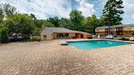 Located at 3500 Pelham Rd in Greenville, South Carolina, this outdoor amenity area is a well-appointed recreational space serving a two-floor residential community. At its centerpiece is a generously sized swimming pool with clear blue water, complemented by an adjoining spa or wading feature. The pool is surrounded by an expansive paver brick patio that provides ample room for lounging and socializing, with multiple chaise lounge chairs positioned to take advantage of the open sky and natural surroundings.

A charming wooden pergola anchors one side of the patio, offering a shaded gathering spot with seating beneath. The area is fully enclosed by a black iron security fence with a gated entry, ensuring privacy and controlled access. A dedicated pool house structure sits adjacent to the pool, featuring an outdoor covered seating area with patio furniture — ideal for relaxing in the shade.

Mature pine trees and lush greenery frame the entire space, creating a private, park-like atmosphere. The two-floor residential buildings with balconies are visible in the backdrop, reinforcing the community setting. The combination of thoughtful landscaping, quality hardscaping, and recreational amenities makes this outdoor space a standout feature of the property.