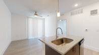This single-floor unit offers a thoughtfully designed open-concept layout that seamlessly connects the kitchen and living area. The kitchen serves as the centerpiece, showcasing a large island with granite countertops, a stainless steel undermount sink, and elegant pendant lighting overhead. Dark wood lower cabinetry is paired with crisp white upper cabinets, while a geometric blue tile backsplash adds a stylish accent. Stainless steel appliances, including a refrigerator, range, and microwave, complete the fully equipped space. Light wood-tone flooring extends throughout, lending warmth and continuity to every room. The open living area benefits from a ceiling fan and a large sliding glass door dressed with vertical blinds, allowing for ample natural light. A dedicated bedroom provides a quiet retreat, featuring its own ceiling fan, a window with blinds, and direct access to the bathroom. The bathroom is finished with granite countertops, white cabinetry, and a tiled tub-shower combination. Recessed lighting and a clean, neutral color palette of white walls and light flooring create a bright, modern ambiance throughout the entire space.