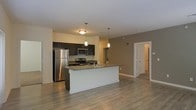 This single-floor space showcases a thoughtfully designed open-concept layout that seamlessly connects the kitchen and living areas. The kitchen is a standout feature, equipped with stainless steel appliances, including a full-sized refrigerator, microwave, and range. Dark espresso cabinetry provides ample storage while adding a bold contrast against the neutral-toned walls. A large central island with a granite countertop serves as both a functional workspace and a casual dining or gathering spot, complete with an integrated sink. Pendant lighting above the island adds a warm, focused glow, complementing the overhead lighting throughout the space. Hardwood-style flooring runs continuously across the open area, enhancing the cohesive and modern aesthetic. The open layout creates a generous sense of space, with clear sightlines from the kitchen through to the living area. A hallway visible in the background leads to additional rooms, suggesting a well-organized floor plan. The neutral color palette of greens and whites throughout contributes to a clean, contemporary ambiance well-suited for modern living.