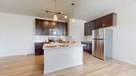 This single-floor residence is designed with an open, flowing layout that seamlessly connects the kitchen and living areas. The kitchen serves as the centerpiece of the space, showcasing rich dark-stained cabinetry with shaker-style doors and brushed nickel hardware. A white quartz-style countertop extends across the perimeter and onto the central island, which features an integrated sink with a modern gooseneck faucet and provides additional seating and prep space. Stainless steel appliances, including a refrigerator, range, and built-in microwave, complement the kitchen's contemporary aesthetic. A white subway tile backsplash adds a clean, classic touch, while pendant lights and a track lighting system provide layered illumination. Light wood-style flooring flows throughout the main living areas, contributing to a bright and airy atmosphere. A window allows natural light into the adjoining living space, enhancing the open feel. The bathroom features a dark-stained vanity with a white countertop, a large mirror, and coordinating light wood flooring. The bedroom is finished with neutral carpet, a ceiling fan, and windows with blinds that invite natural light.