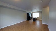 This three-floor space showcases a thoughtfully designed open-concept main living area that seamlessly integrates the kitchen and living room. The kitchen is a standout feature, anchored by a dark-toned island with a double stainless steel sink and rich granite-style countertops. Dark cabinetry complements a suite of black appliances, including a French door refrigerator and range, creating a bold, cohesive aesthetic. Recessed lighting is distributed across the ceiling, providing consistent, bright illumination throughout the open area. The living space is generously proportioned, offering ample room for a variety of furniture arrangements. Warm hardwood-style flooring flows continuously throughout, adding a welcoming feel underfoot. Neutral-toned walls contribute to a clean, versatile backdrop suited to many décor styles. A window along one wall allows natural light to filter in, brightening the space. Visible HVAC vents indicate a central air system for year-round comfort. The overall design balances modern finishes with functional layout across all three floors of the property.