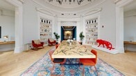 Located at 901 Second Ave N in Nashville, Tennessee, this single-floor space showcases an exceptional blend of historic architectural grandeur and contemporary interior design. The centerpiece of the space is a striking rotunda room distinguished by a soaring domed ceiling adorned with elaborate white plasterwork moldings and a bold black chandelier. Built-in bookshelves line the curved walls, lending a library-like sophistication to the room. A dramatic live-edge wood conference table with intricate dark vein detailing commands the center of the space, surrounded by vibrant orange and red chairs atop a richly colored, patterned area rug. Comfortable red rocking chairs and accent seating complete the rotunda, while curated art pieces—including a vivid red sculptural figure—add eclectic flair. An adjacent lounge area connects seamlessly through an arched opening, offering additional flexible seating. The entry area contrasts beautifully with its modern aesthetic, featuring floor-to-ceiling black-framed glass doors and windows that flood the herringbone hardwood floors with natural light and provide open street views. Together, these spaces create an atmosphere that is both architecturally distinguished and thoughtfully curated.