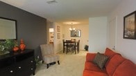 This single-floor residence offers an open-concept layout that seamlessly connects the living and dining areas, creating a cohesive and functional flow throughout the space. The living area features warm, neutral-toned carpeting that extends throughout, adding comfort underfoot, while a bold dark accent wall provides a striking contrast against the lighter surrounding walls, lending the space a modern and stylish character. A media console anchors one corner of the living area, complemented by accent seating that adds a touch of classic charm. The dining area is a standout feature, centered around a dark wood dining table and chair set positioned beneath an elegant multi-arm chandelier that casts warm, inviting light. A decorative mirror on the dining room wall enhances the sense of depth and reflects the warm ambiance of the room. Framed artwork adorns the walls, adding a personalized touch. A glimpse into the adjacent kitchen reveals cabinetry and countertops, suggesting a practical and connected cooking space. The overall atmosphere is warm, welcoming, and thoughtfully designed, with a neutral palette punctuated by rich tones that create a balanced and comfortable living environment.