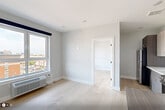 Unit 502 at 1213–1221 Broad St is a well-designed one-bedroom, one-bathroom residence that makes efficient use of its compact footprint. The open layout is anchored by light-toned hardwood flooring that runs throughout, lending warmth and visual continuity to the space.

The kitchen is a standout feature, outfitted with modern two-tone cabinetry in warm gray and taupe finishes, a white subway tile backsplash, stainless steel appliances including a range, microwave, and dishwasher, and a peninsula with a dark countertop that provides additional prep and dining space.

The living area flows naturally from the kitchen and benefits from large windows dressed with navy roller shades, allowing for generous natural light and elevated views of the surrounding cityscape. A wall-mounted HVAC unit ensures year-round climate control.

The bedroom is a comfortable, well-proportioned room with the same light hardwood flooring, large windows offering urban views, and clean white walls that create a calm, neutral atmosphere.

The bathroom is finished with neutral large-format tile on both the floors and shower surround, complemented by matte black fixtures and hardware, including grab bars, lending a sleek and modern aesthetic.

Overall, the unit presents a clean, contemporary design with consistent finishes throughout, making it a functional and stylish urban residence.