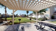 This single-level outdoor community amenity space is thoughtfully designed to offer residents a comprehensive recreational environment. At its centerpiece is a well-maintained swimming pool, enclosed by a secure wrought iron fence and surrounded by a generous concrete pool deck furnished with lounge chairs and shaded patio seating beneath green umbrellas — ideal for relaxing on warm, sunny days. Palm trees and mature landscaping frame the pool area, adding a resort-like ambiance under open blue skies.

Adjacent to the pool, a substantial white pergola structure provides shaded outdoor seating and picnic areas complete with metal picnic tables and benches — a functional space for social gatherings and outdoor dining. Nearby BBQ grills are conveniently positioned to support outdoor cooking. A well-kept lawn area with neatly trimmed shrubbery and walkways connects the various amenity zones, creating a cohesive and inviting outdoor environment.

Access to the covered clubhouse or amenity building is provided through elegant white-framed French doors set into a stucco exterior wall, which also features an emergency shut-off switch panel. The surrounding residential buildings are visible beyond the fenced perimeter, reinforcing the sense of a close-knit community. The overall atmosphere is open, bright, and welcoming throughout.