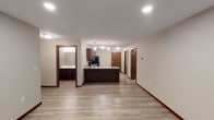 This single-floor space offers a clean, modern design with an open-concept layout that connects the main living area to the kitchen seamlessly. The kitchen is a standout feature, anchored by a substantial island with a stone-look countertop and rich, dark wood cabinetry that provides ample storage. Pendant lighting hangs above the island, adding a refined touch, while recessed ceiling lights illuminate the space throughout. Stainless steel appliances complement the contemporary aesthetic. Light wood-look plank flooring extends across the main area, creating a warm and cohesive feel that contrasts elegantly with the dark wood trim and cabinetry. Adjacent to the kitchen, a hallway leads to multiple doors, suggesting well-defined private rooms beyond. A bathroom is also visible off the main space, featuring a vanity with dark wood cabinetry and a mirror, finished with coordinating tile flooring. The neutral wall tones and recessed lighting contribute to a bright, airy ambiance throughout. Overall, the space presents a thoughtfully designed interior that balances functional layout with modern, cohesive finishes.