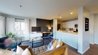 Located at 320 SW Montgomery Street in Portland, Oregon, this well-appointed apartment on the third floor offers a thoughtfully designed single-level layout with modern finishes throughout. The open-concept living area is anchored by large windows dressed with sheer white curtains, flooding the space with natural light and offering views of the surrounding neighborhood. Dark hardwood-style flooring flows throughout, complementing a bold charcoal accent wall that adds depth and contrast to the living room. A dedicated workspace with a wooden desk sits seamlessly within the living area, making the layout highly functional. The kitchen features white cabinetry, a white subway tile backsplash, light stone countertops, stainless steel appliances, and a peninsula with a built-in sink — ideal for both cooking and casual entertaining. The bedroom is a serene retreat, highlighted by a dramatic dark accent wall, a decorative oversized floral art piece, and a cozy, well-styled layout. The bathroom includes a clean white vanity with ample storage, a framed mirror, and a shower with a stylish curtain. Throughout the unit, the design balances contemporary aesthetics with functional comfort, creating a cohesive and inviting urban living environment.