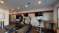 Located at 1001 Toms Creek Rd in Blacksburg, VA, this fitness center is a thoughtfully designed, single-floor facility built to support a wide range of workout routines. The space features durable rubber flooring throughout, recessed lighting, and a bold red accent stripe running along the upper walls, giving the room a dynamic, energetic atmosphere.

Cardio equipment is well-represented, with a curved treadmill, stationary bikes — including a Peloton-style connected cycle — and additional aerobic machines. A mounted TRX training station provides options for suspension-based functional fitness, while a wall-mounted medicine ball rack holds a colorful assortment of weighted balls. BOSU balance trainers and kettlebells round out the functional training area. A dedicated dumbbell rack ensures strength training needs are met.

Multiple flat-screen televisions are mounted on the walls, keeping users entertained during workouts. A water fountain station is conveniently positioned near the entrance, and motivational framing — including quotes such as "if it doesn't challenge you, it doesn't change you" — adds an inspiring touch to the environment.

Natural light enters through windows fitted with blinds, and glass double doors provide direct outdoor access, connecting the interior to the surrounding community grounds. The overall design is clean, functional, and purpose-driven.