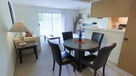 This single-floor residence is designed around an open-concept layout that seamlessly connects the kitchen, dining area, and living room, creating a cohesive and functional living environment. The kitchen features light wood cabinetry, a white countertop peninsula that doubles as a breakfast bar, and standard appliances including a full-size refrigerator. Track lighting overhead enhances visibility throughout the cooking and dining areas. Adjacent to the kitchen, the dining space accommodates a round dark-wood table with matching leather chairs, providing an intimate setting for meals. A dedicated laundry area is visible just off the kitchen, adding everyday convenience. The living room is carpeted and comfortably proportioned, anchored by a large sliding glass door that bathes the space in natural light and opens to an outdoor patio. The view from this door is a notable highlight, overlooking a tranquil pond framed by lush greenery and neighboring buildings. The neutral color palette throughout — soft walls, light flooring, and understated finishes — contributes to a calm, welcoming ambiance suited for comfortable everyday living.