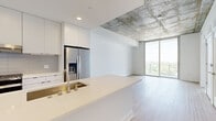 This contemporary single-floor residence showcases a refined industrial-modern aesthetic throughout. The open-concept layout is anchored by an exposed concrete ceiling that contrasts beautifully with crisp white walls and light-toned hardwood flooring, creating a sophisticated urban atmosphere.

The kitchen is a standout feature, centered around a generous white quartz island with an undermount sink and brushed gold fixtures. Flat-front white cabinetry extends to the ceiling, complemented by a subway tile backsplash and a full suite of stainless steel appliances, including a French door refrigerator with ice dispenser and a gas range.

The living area opens directly to a balcony through floor-to-ceiling sliding glass doors, framing sweeping views of the city skyline and surrounding tree canopy. Additional rooms benefit from large gridded windows that flood the space with natural light while offering elevated views of the cityscape.

Both bathrooms feature a consistent design language: white flat-front vanities with quartz countertops, undermount sinks with brushed nickel fixtures, frameless mirrors flanked by globe sconce lighting, and distinctive patterned tile flooring with a geometric star motif. One bathroom includes a glass-enclosed walk-in shower with white subway tile surround.