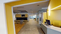 This well-appointed single-floor amenity center is designed with a vibrant, contemporary aesthetic, anchored by a striking yellow and grey color scheme that carries throughout every zone. At the heart of the space is a spacious lounge area featuring a large sectional sofa surrounding a sleek linear fireplace, complemented by stylish swivel chairs and a geometric-patterned carpet that adds visual depth. Large floor-to-ceiling windows along the perimeter flood the space with natural light and offer views of the surrounding streetscape, while a circular curved seating nook near the entrance provides an inviting spot to relax. A dedicated dining area features warm hardwood flooring, bistro-style tables and chairs, pendant lighting in a cylindrical yellow design, and curated artwork displayed along the walls. Adjacent to the dining zone is a kitchenette equipped with a long white bar counter, bold yellow barstools, stainless steel appliances, and a wall-mounted television. The computer workstation area offers multiple desktops set against a textured grey accent wall. A fully equipped fitness center, visible through a wide open archway, houses cardio and strength training equipment. A foosball table and additional recreational elements round out the game area, making this a comprehensive, community-focused facility.