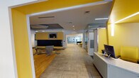 This well-appointed single-floor amenity center is designed with a vibrant, contemporary aesthetic, anchored by a striking yellow and grey color scheme that carries throughout every zone. At the heart of the space is a spacious lounge area featuring a large sectional sofa surrounding a sleek linear fireplace, complemented by stylish swivel chairs and a geometric-patterned carpet that adds visual depth. Large floor-to-ceiling windows along the perimeter flood the space with natural light and offer views of the surrounding streetscape, while a circular curved seating nook near the entrance provides an inviting spot to relax. A dedicated dining area features warm hardwood flooring, bistro-style tables and chairs, pendant lighting in a cylindrical yellow design, and curated artwork displayed along the walls. Adjacent to the dining zone is a kitchenette equipped with a long white bar counter, bold yellow barstools, stainless steel appliances, and a wall-mounted television. The computer workstation area offers multiple desktops set against a textured grey accent wall. A fully equipped fitness center, visible through a wide open archway, houses cardio and strength training equipment. A foosball table and additional recreational elements round out the game area, making this a comprehensive, community-focused facility.